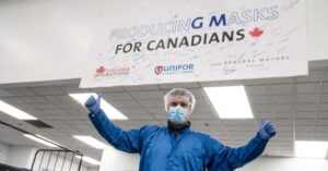 Employee at Oshawa Operations proudly stands under a sign reading "Producing Masks for Canadians," showcasing a cooperative initiative with Unifor and General Motors to manufacture masks.