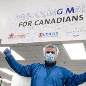 Man in protective gear stands under a large sign that reads "Producing Masks for Canadians" in a facility, showcasing support from Unifor and demonstrating commitment to manufacturing safety equipment.