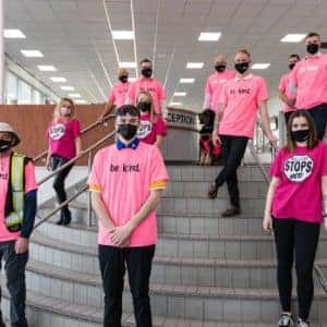 Group of diverse individuals wearing bright pink shirts and black masks, standing on stairs in a public building to promote kindness and support a positive message.