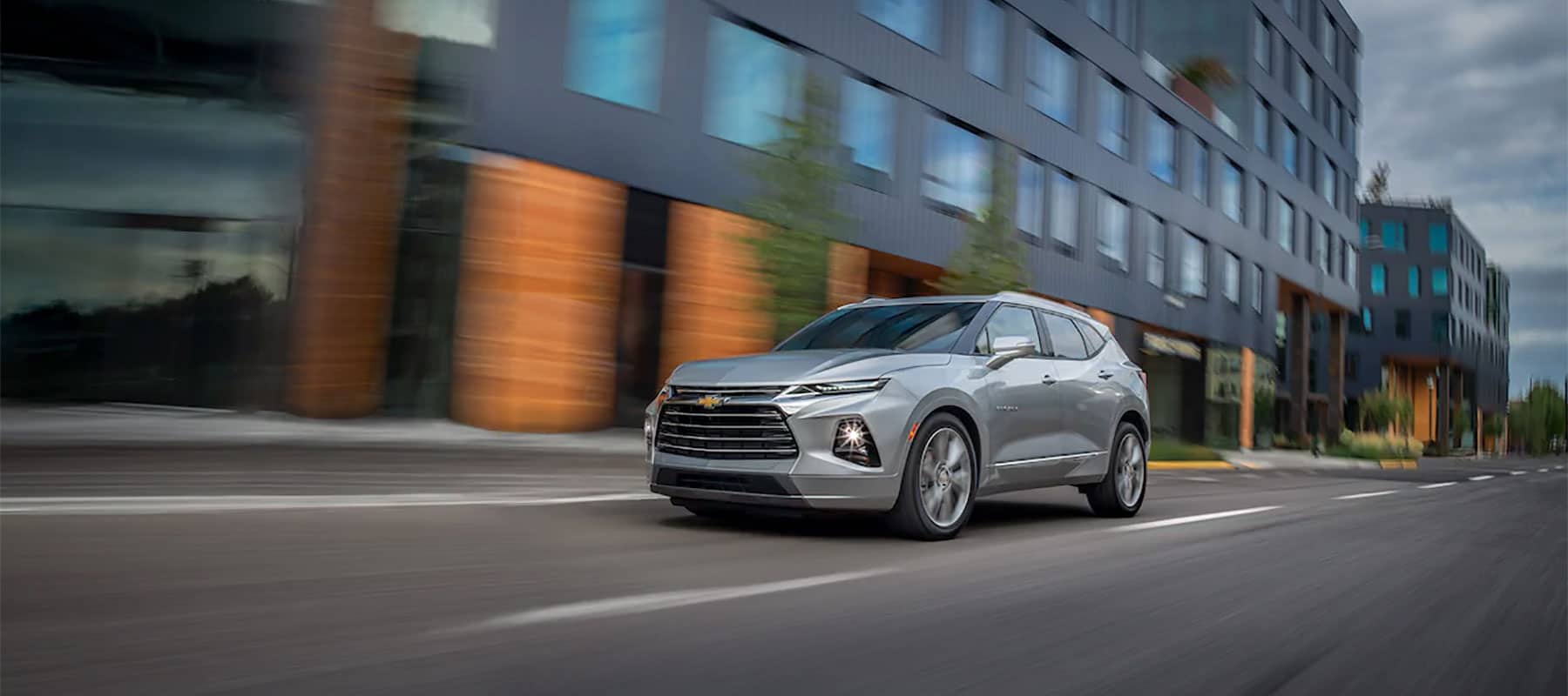 Silver Chevrolet Blazer driving on a city street with modern buildings in the background, showcasing its sleek design and dynamic performance.