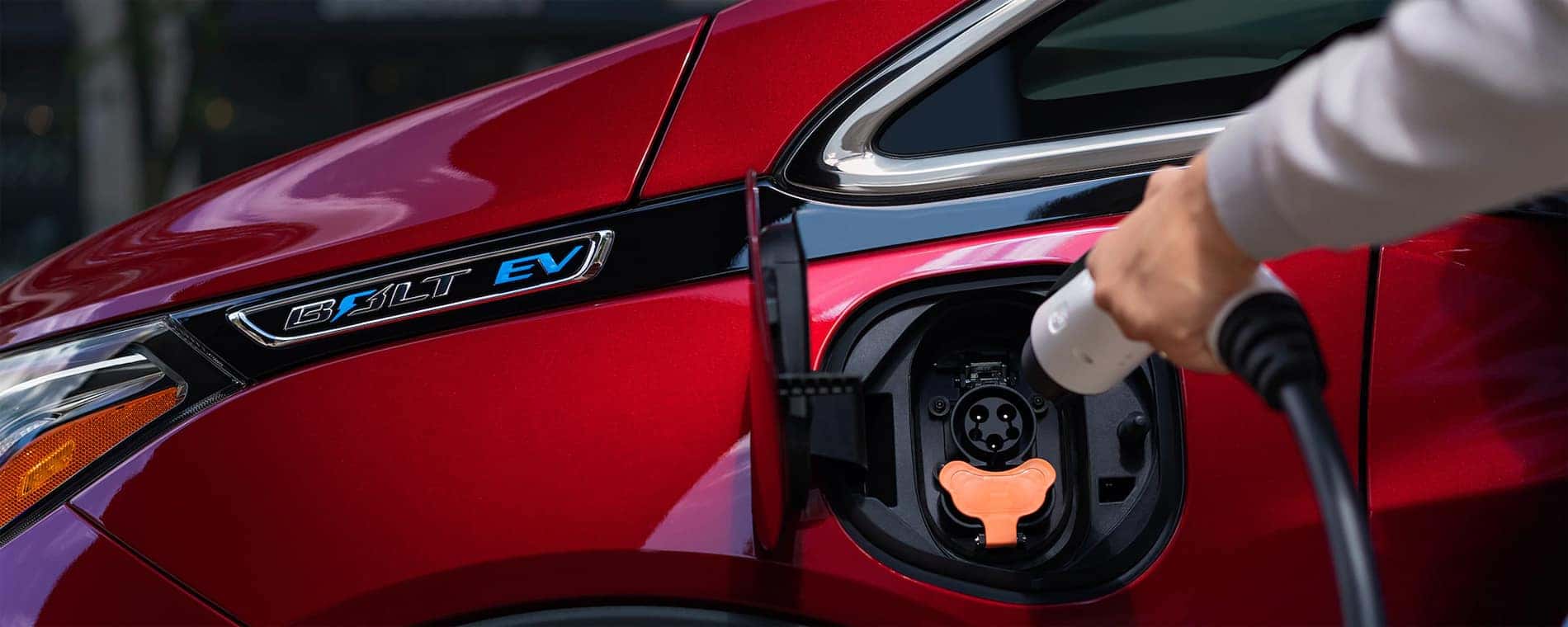 Close-up of a red Chevrolet Bolt EV electric vehicle charging, showcasing the charging port with a hand inserting a charging connector. The image highlights the modern design and eco-friendly features of electric cars.