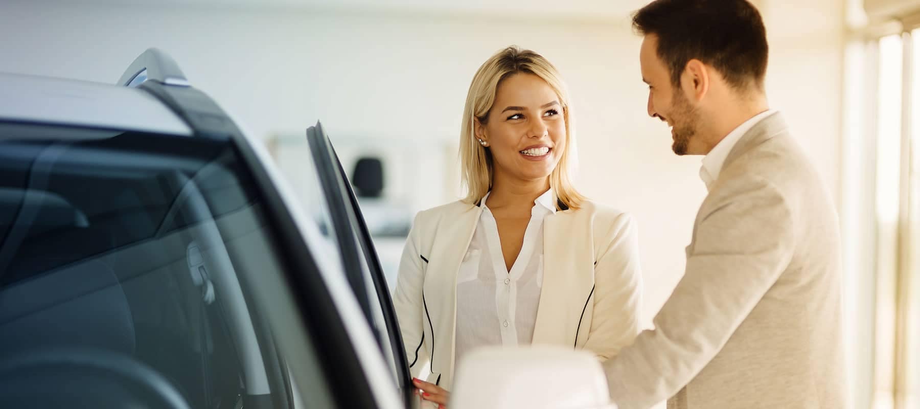 Smiling woman in a white blazer discussing car features with a man in a light gray suit at a car dealership, emphasizing customer service and automotive sales.