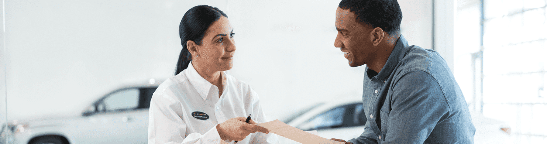Customer service interaction at a car dealership, featuring a smiling woman in a white shirt handing a document to a man in a blue shirt, both engaged in a friendly conversation.