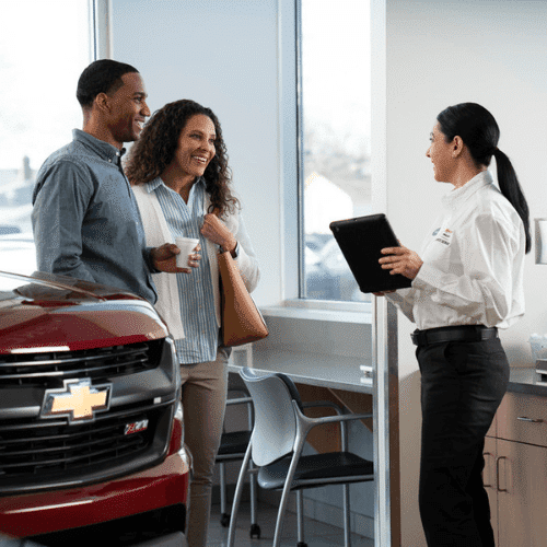 Couple interacting with a dealership representative inside a car showroom, discussing vehicle services while a Chevrolet vehicle is prominently displayed in the foreground.