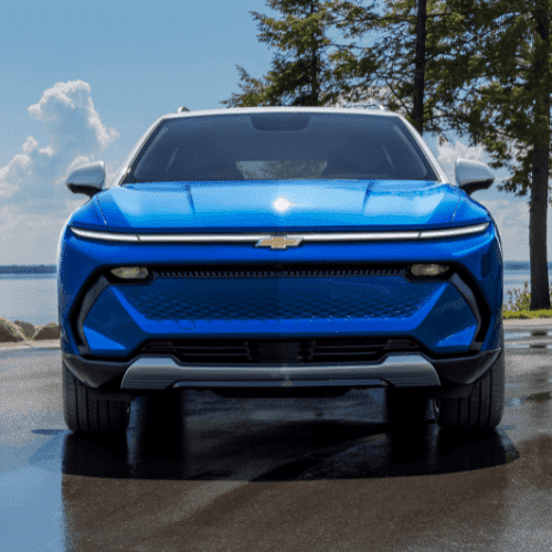 Front view of a blue Chevrolet vehicle parked by a lake, showcasing a sleek design with a prominent logo and modern features, surrounded by trees and a clear sky.