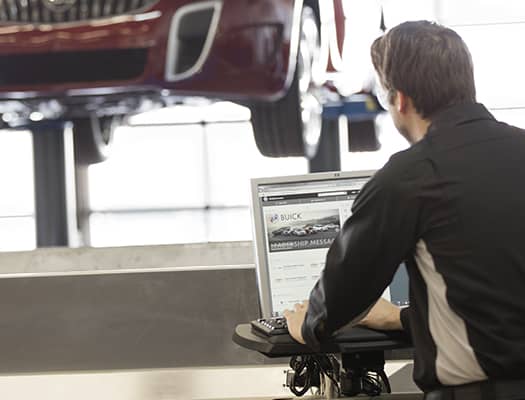 Automotive technician working on a computer in a service bay with a Buick vehicle on a lift, focusing on vehicle maintenance and online service scheduling.