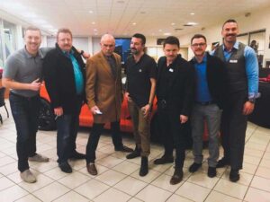Group of seven men posing together in a car dealership showroom, with an orange car in the background. The men wear a mix of business casual and formal attire, showcasing camaraderie and professionalism in a retail environment.