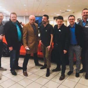 Group of professional men posing together in a car showroom, with an orange vehicle in the background. The setting features a modern interior suitable for automotive exhibitions or events.