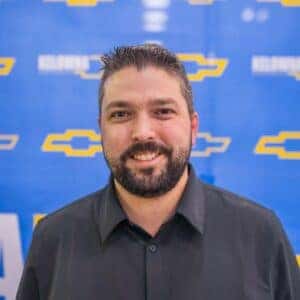 Smiling man in a gray sweater standing in front of a blue Chevrolet-branded backdrop, promoting automotive services.