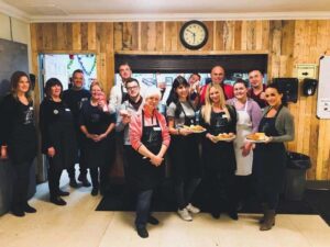 A group of volunteers smiling and posing in a community kitchen, wearing aprons and holding plates of food, showcasing teamwork and hospitality in a warm, inviting environment.