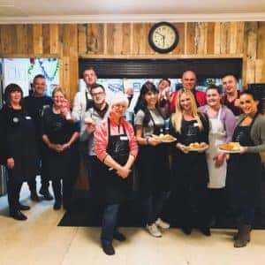 Group of diverse individuals wearing aprons, standing together in a cozy kitchen environment, smiling and holding freshly baked goods, showcasing teamwork and community involvement in cooking activities.