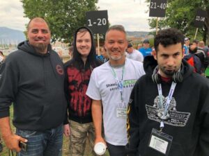 Group of four men smiling at an outdoor event, standing in front of numbered signs. The setting features trees and a scenic background, with attendees visible in the distance, suggesting a community gathering or race event. Each man wears casual clothing, and some have event lanyards around their necks.