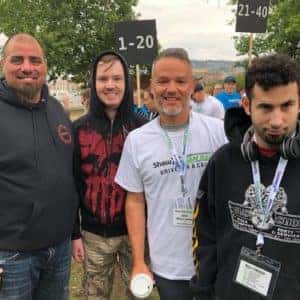 Group of four men at an outdoor event, smiling and posing together, with numbered signs in the background indicating different categories. They appear to be participating in a friendly gathering or competition, showcasing camaraderie and community spirit.
