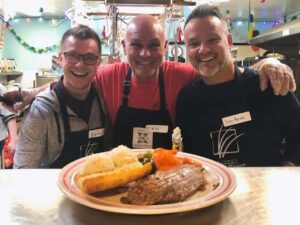 Three smiling volunteers pose in a kitchen, proudly showcasing a plated meal featuring meat, mashed potatoes, and vegetables. The men wear aprons with name tags and contribute to a community food initiative. Decorated walls and festive lights create a cheerful atmosphere.