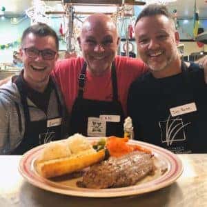 Three smiling chefs wearing aprons pose together in a kitchen setting, showcasing a plate of freshly prepared meatloaf, mashed potatoes, and vegetables. The atmosphere is warm and inviting, reflecting a community cooking event.