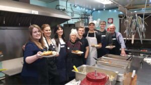 Group of eight volunteers in a kitchen setting, wearing aprons and holding plates of food, with a well-equipped cooking area in the background and various kitchen utensils hanging above.