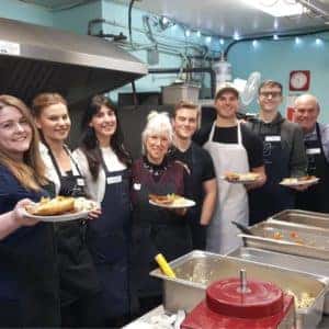 Group of volunteers in a kitchen holding plates of food, wearing aprons and name tags, engaged in food service or community outreach.