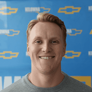 Smiling man in a gray sweater standing in front of a blue Chevrolet-branded backdrop, promoting automotive services.