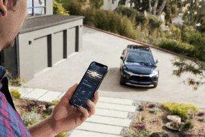 Person using a smartphone to control a smart car in a driveway surrounded by greenery, showcasing modern technology and convenience in a residential setting.