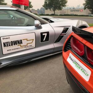 Silver Chevrolet Corvette with a promotional sign for Kelowna Chevrolet, featuring the number 7, parked alongside an orange Corvette with a logo for the Okanagan Dream Rally, set against a hazy background.