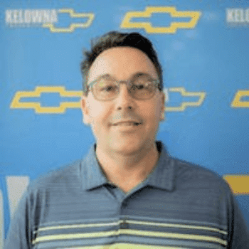 Smiling man in a gray sweater standing in front of a blue Chevrolet-branded backdrop, promoting automotive services.