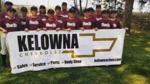 Youth baseball team from Kelowna posing with a banner featuring the Kelowna Chevrolet logo, promoting sales, service, parts, and body shop services. Team members wear matching maroon jerseys and caps, standing outdoors in a grassy area.