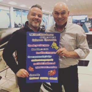 Two smiling men hold a brightly colored promotional poster in a retail setting, showcasing various products and information related to a business event. The background features modern store decor, emphasizing a community-oriented atmosphere.