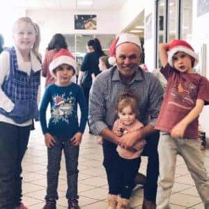 A group of five individuals, including four children wearing Santa hats and an adult man, smiling and posing together in a festive atmosphere. The setting appears to be a community center or event hall decorated for the holiday season.