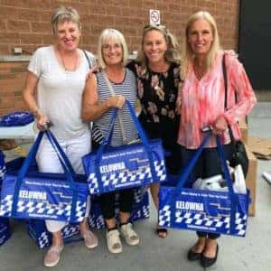 Four women smiling and posing together while holding blue tote bags marked "Kelowna" against a brick wall background, showcasing a community event or donation initiative.