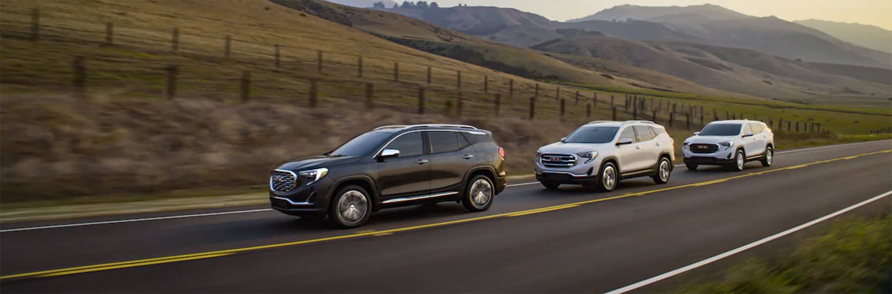 Three GMC SUVs driving on a scenic highway surrounded by rolling hills, showcasing their sleek designs and modern features.