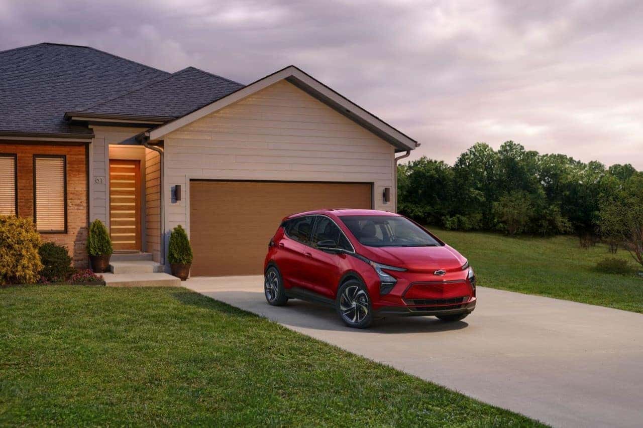 Red electric car parked in front of a modern house with a landscaped yard and evening sky, showcasing a stylish driveway and contemporary architecture.