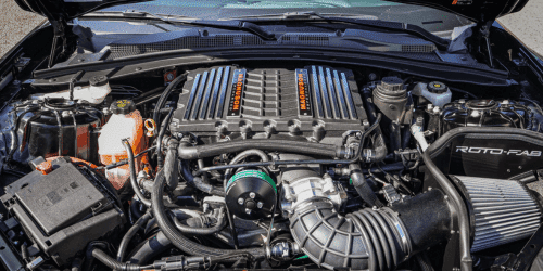 Close-up view of a performance car engine, showcasing a supercharger, Roto-Fab cold air intake, and various components including pipes, tanks, and wiring, all under the hood of a high-performance vehicle.