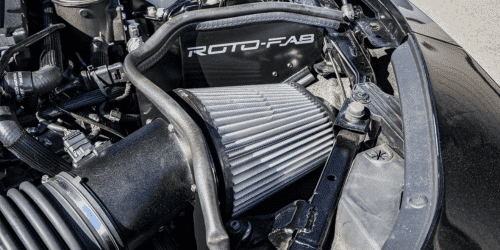 Close-up view of a car engine highlighting a Roto-Fab air intake system, featuring a polished aluminum filter and surrounding components, designed to enhance engine performance and airflow efficiency.
