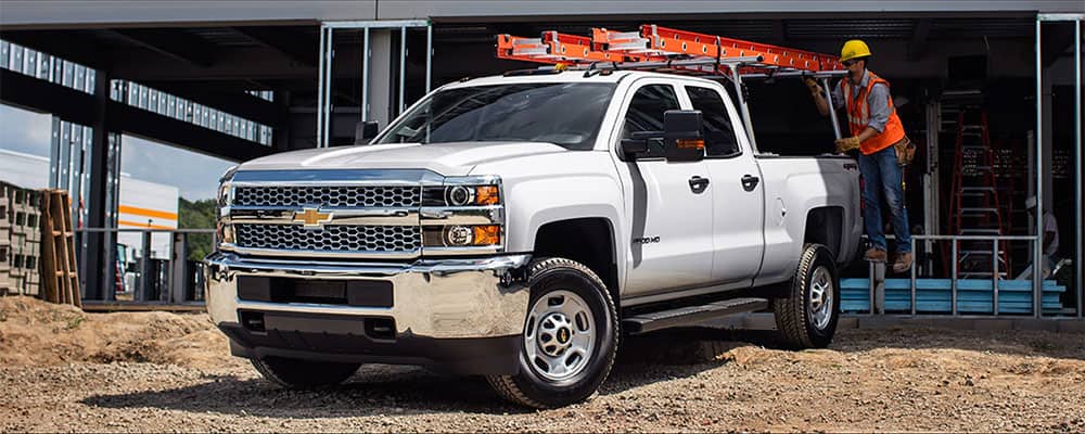 Construction site featuring four Chevrolet trucks parked on sandy ground, with workers in safety gear attending to equipment. A partially constructed building is visible in the background.