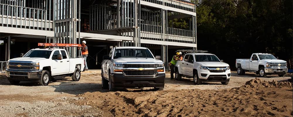 Construction site featuring four Chevrolet trucks parked on sandy ground, with workers in safety gear attending to equipment. A partially constructed building is visible in the background.