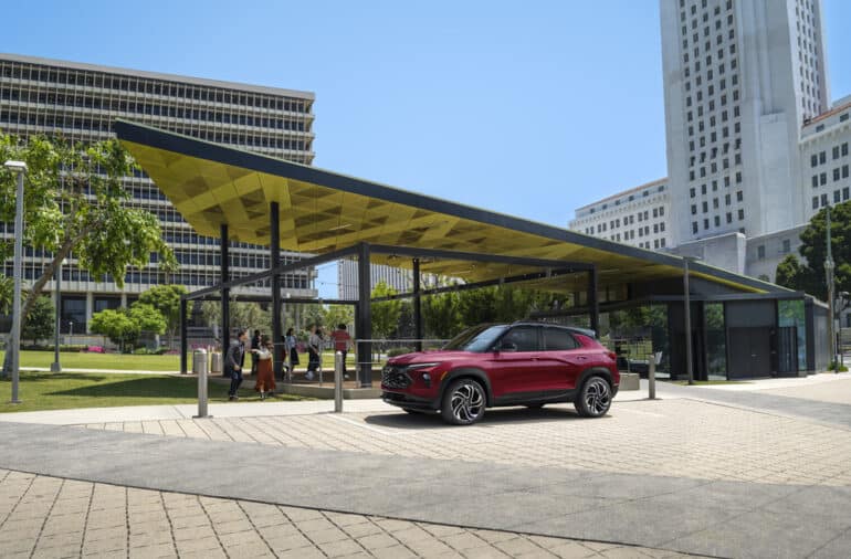 Red SUV parked in front of a modern pavilion with a green space and tall buildings in the background, showcasing an urban park setting on a sunny day.