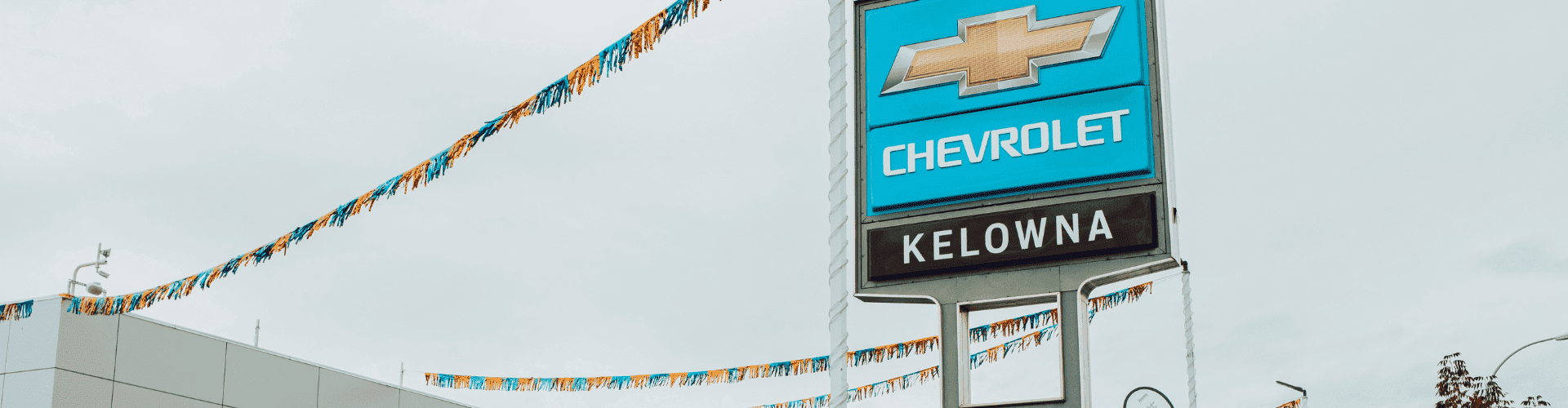 Chevrolet dealership sign in Kelowna, featuring a large blue logo and colorful banners overhead against a cloudy sky.