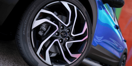 Close-up image of a modern car wheel featuring a stylish multi-spoke design in black and silver, mounted on a performance tire. The backdrop showcases a vibrant and colorful environment, emphasizing the vehicle's sporty aesthetic.