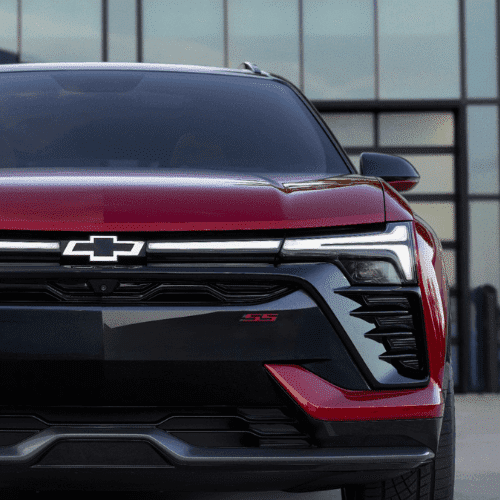 Side view of a sleek red SUV parked in front of a modern garage with large glass doors, showcasing its aerodynamic design and stylish alloy wheels. Ideal for automotive enthusiasts looking for a blend of luxury and performance.