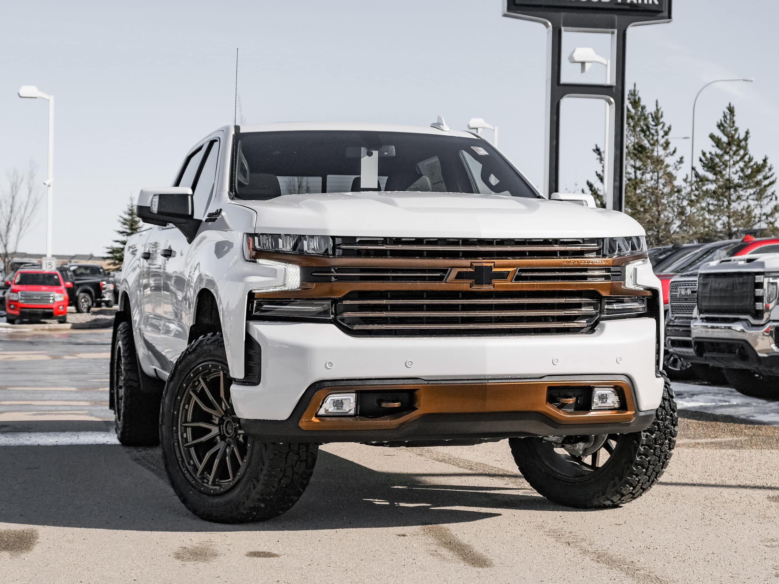 Front view of a white Chevrolet Silverado with bronze accents, showcasing its rugged design and large all-terrain tires, parked in a dealership lot. Other trucks are visible in the background, highlighting the truck's dominant presence.