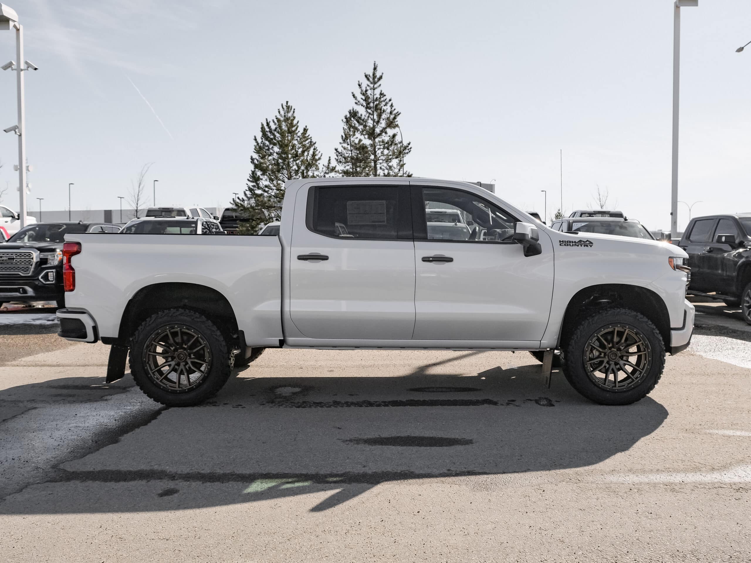 Close-up of a black pickup truck showcasing custom oversized bronze rims and rugged tires, parked on a paved road with a clear sky in the background. The vehicle's sleek profile features a prominent side mirror and modern design elements.