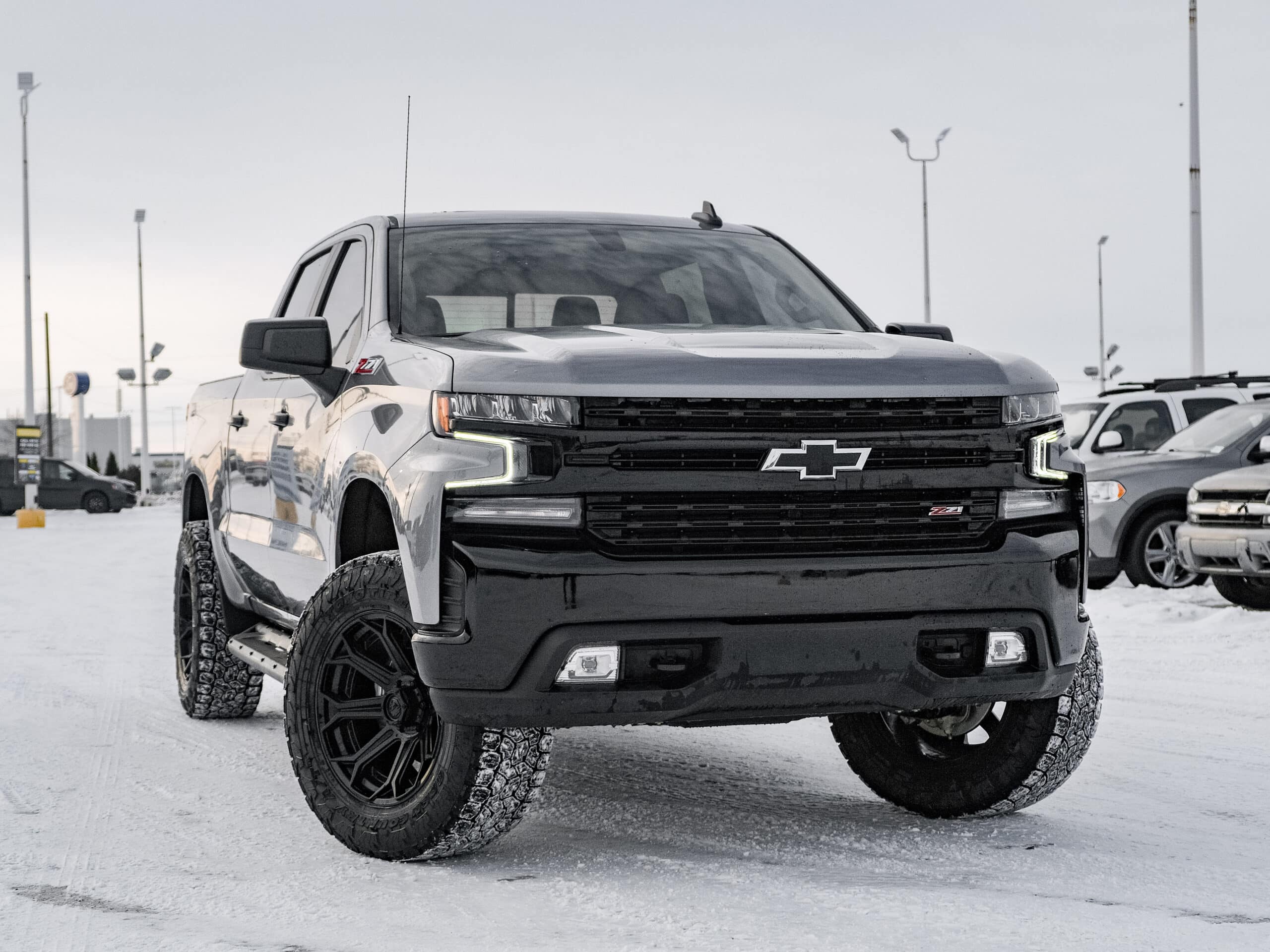 Black Chevrolet Silverado pickup truck parked on snow-covered ground, showcasing rugged tires and modern design, with dealership in the background. Ideal for off-road enthusiasts and winter driving conditions.