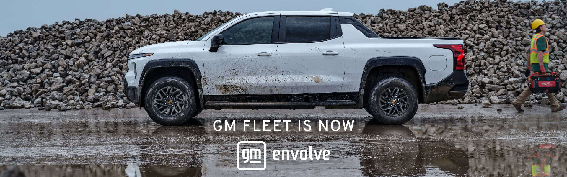 White pickup truck parked on a construction site with a worker carrying tools, featuring a backdrop of a rock pile. Text overlay reads "GM Fleet is Now Envolve."
