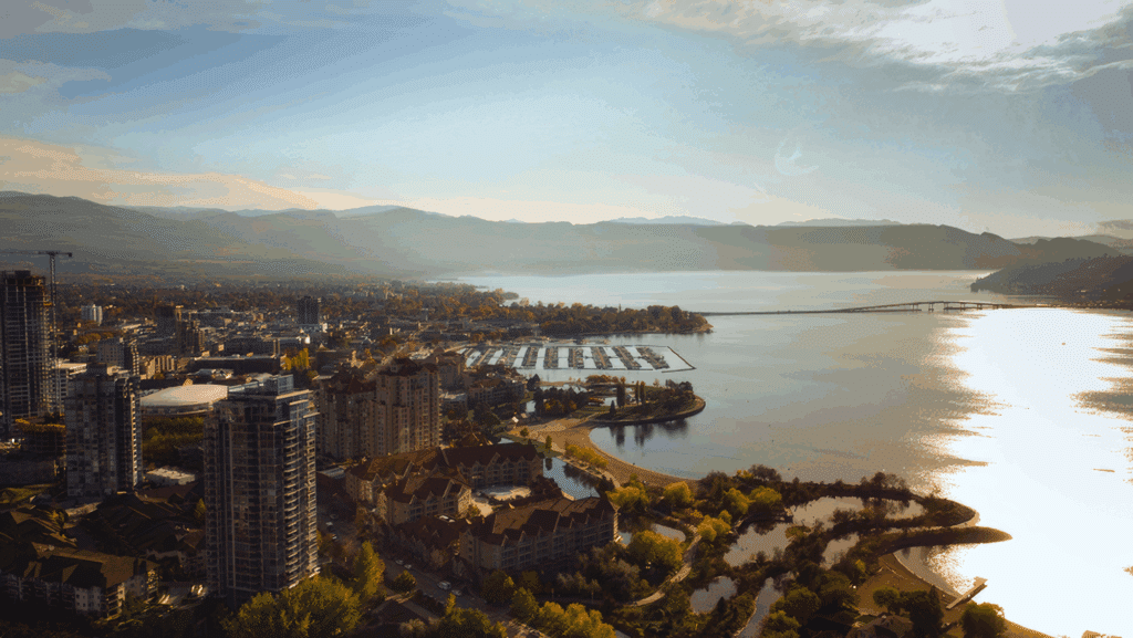 Aerial view of a lakeside cityscape featuring modern high-rise buildings, a marina with docked boats, and scenic waterfront parks along a serene lake, framed by mountains under a clear blue sky.