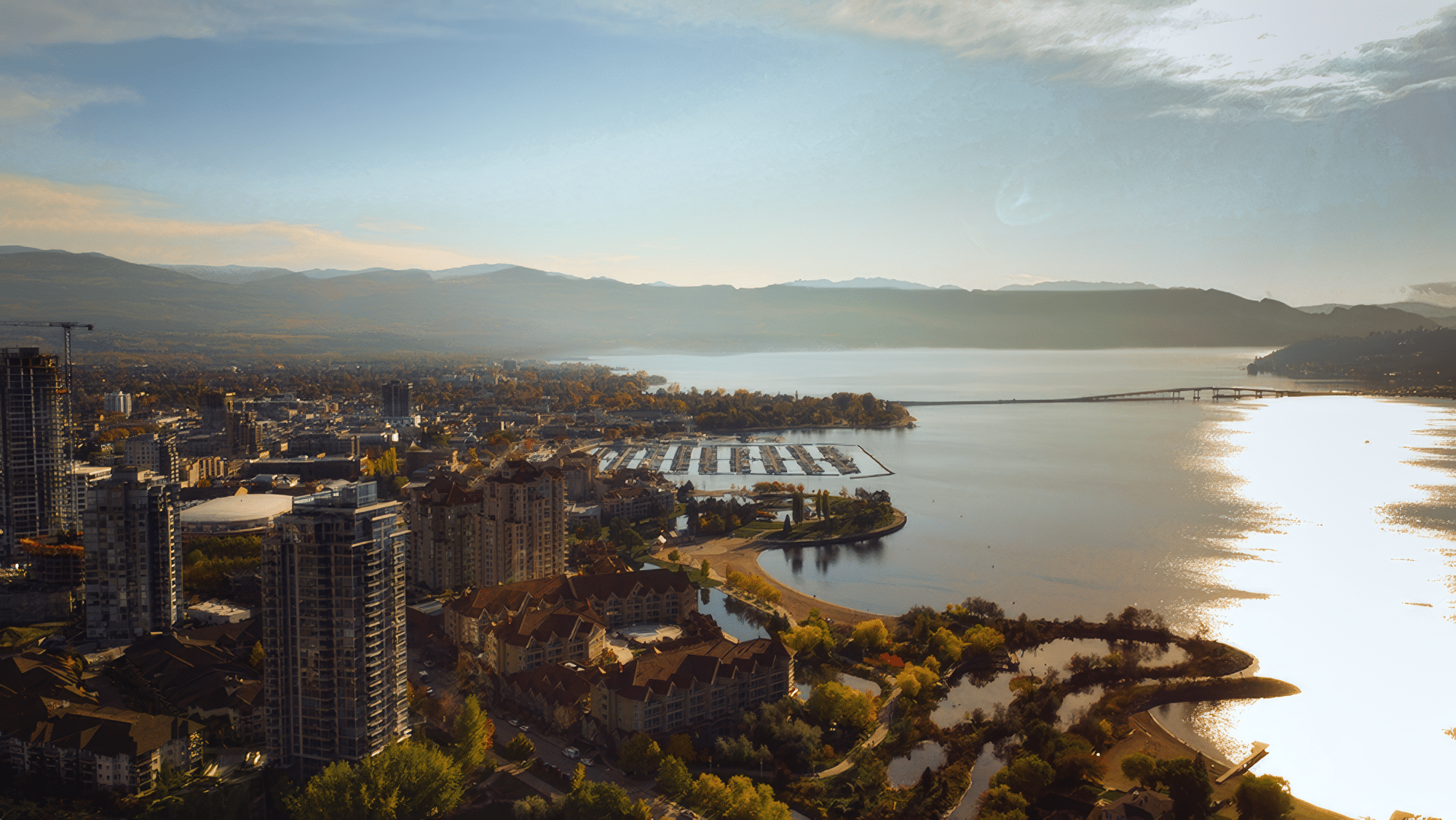 Aerial view of a lakeside cityscape featuring modern high-rise buildings, a marina with docked boats, and scenic waterfront parks along a serene lake, framed by mountains under a clear blue sky.