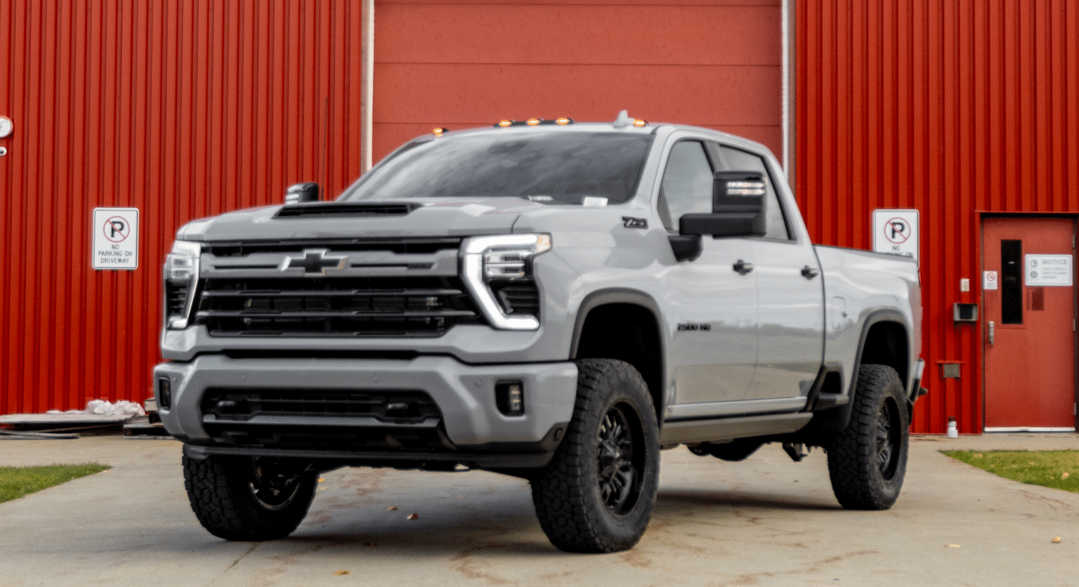 2023 Chevrolet Silverado 2500HD in metallic gray parked in front of a red building, showcasing rugged design and off-road capabilities with all-terrain tires and LED lights.
